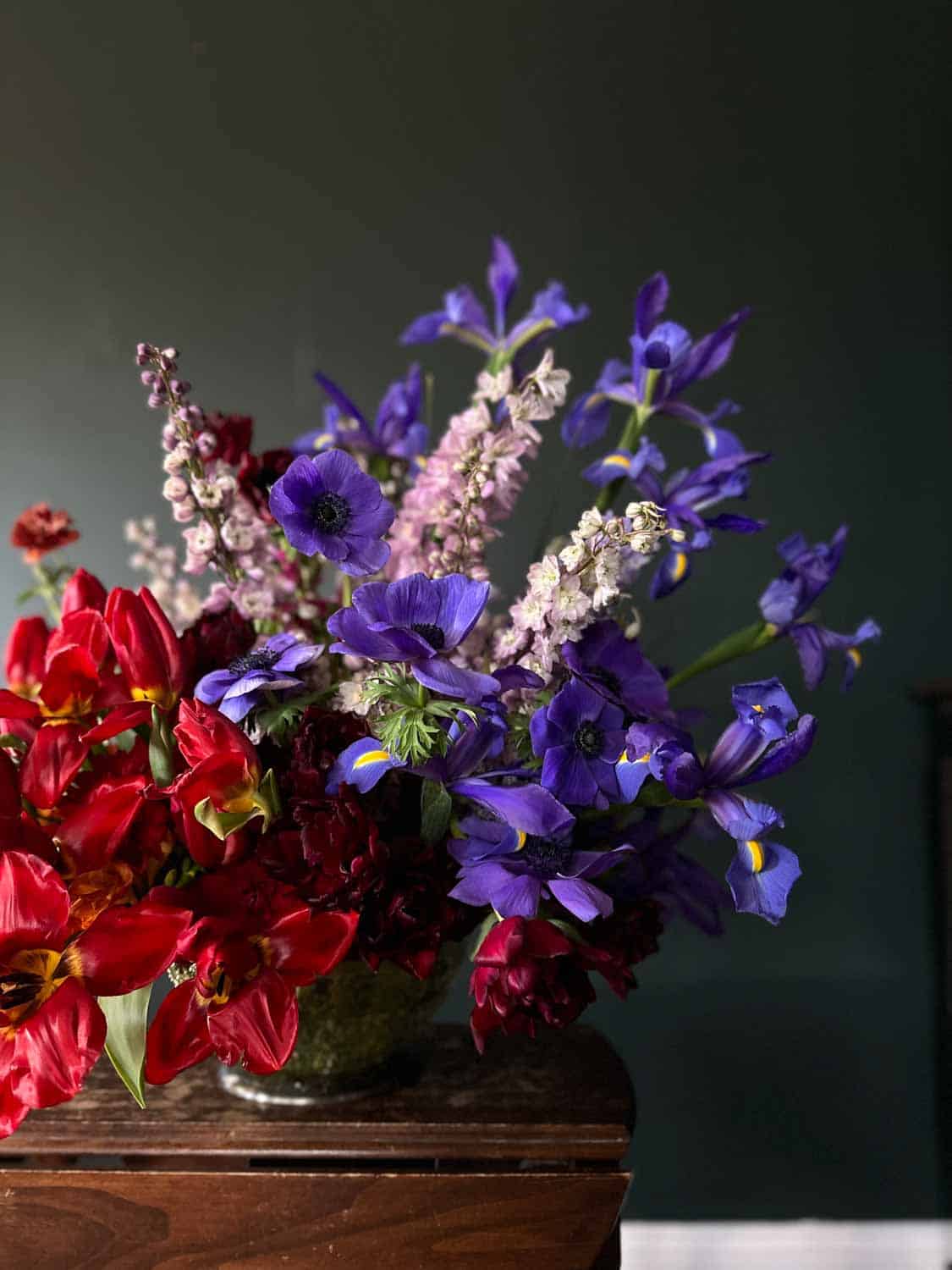 A bouquet of red, pink, and purple flowers arranged in a vase on a wooden table against a dark background.