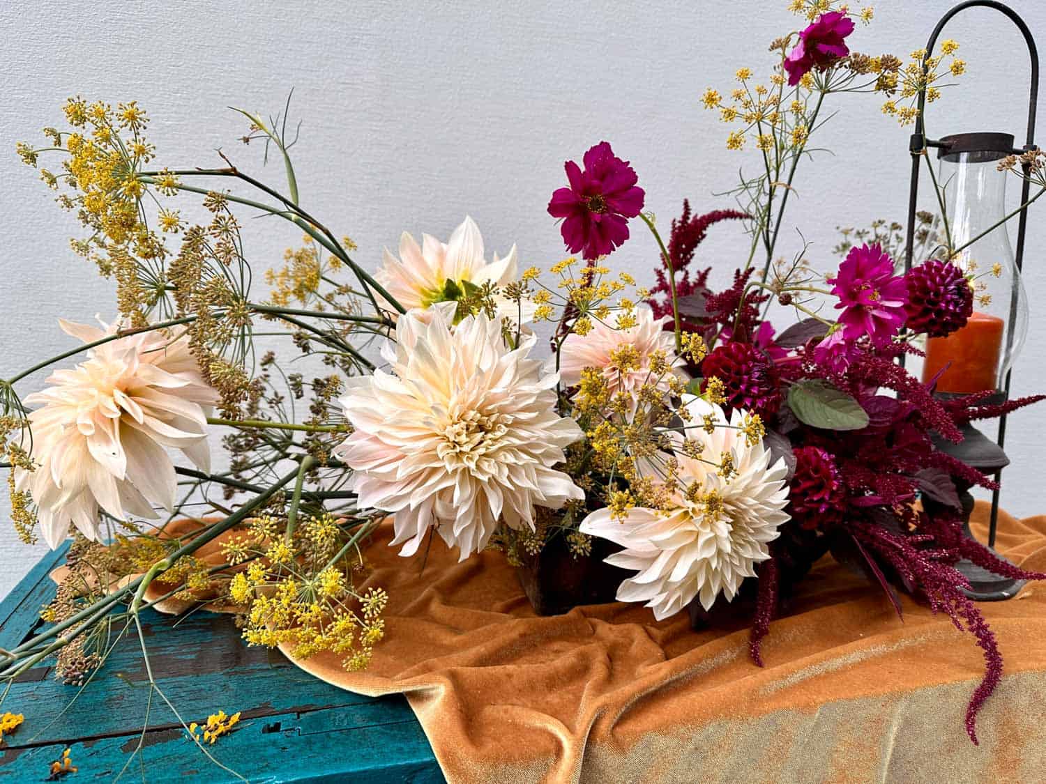 A floral arrangement with white and pink flowers and green foliage on an orange cloth, next to a black lantern and candle.