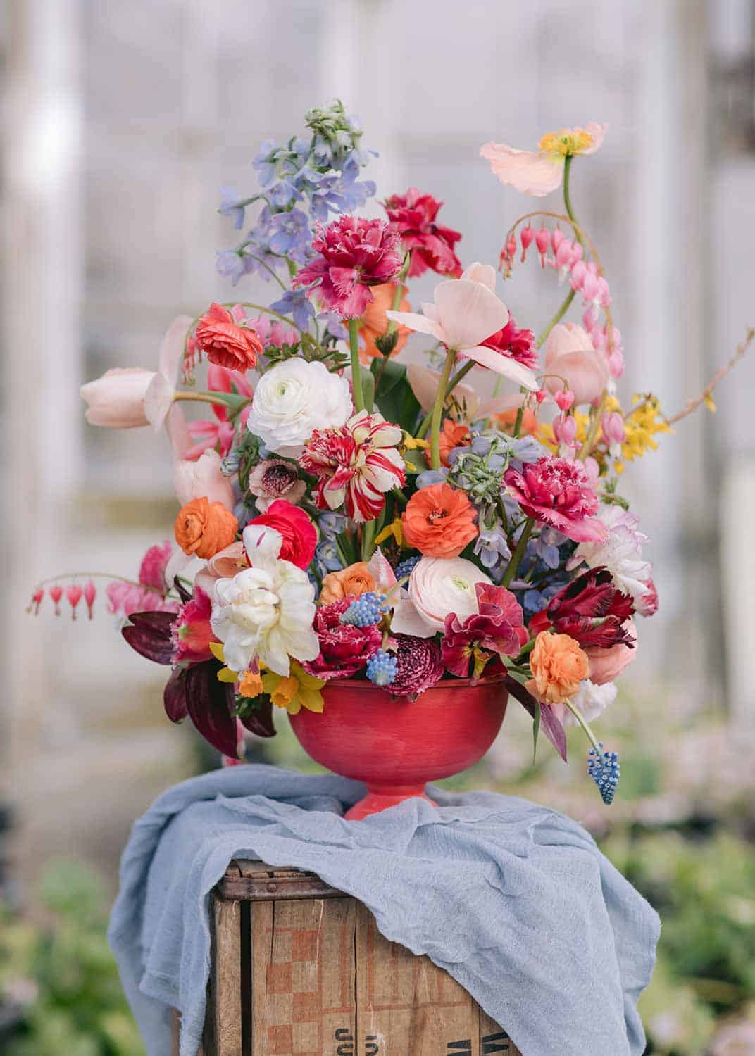 A vibrant floral arrangement with assorted flowers in a red vase, placed on a wooden crate draped with a light blue cloth.