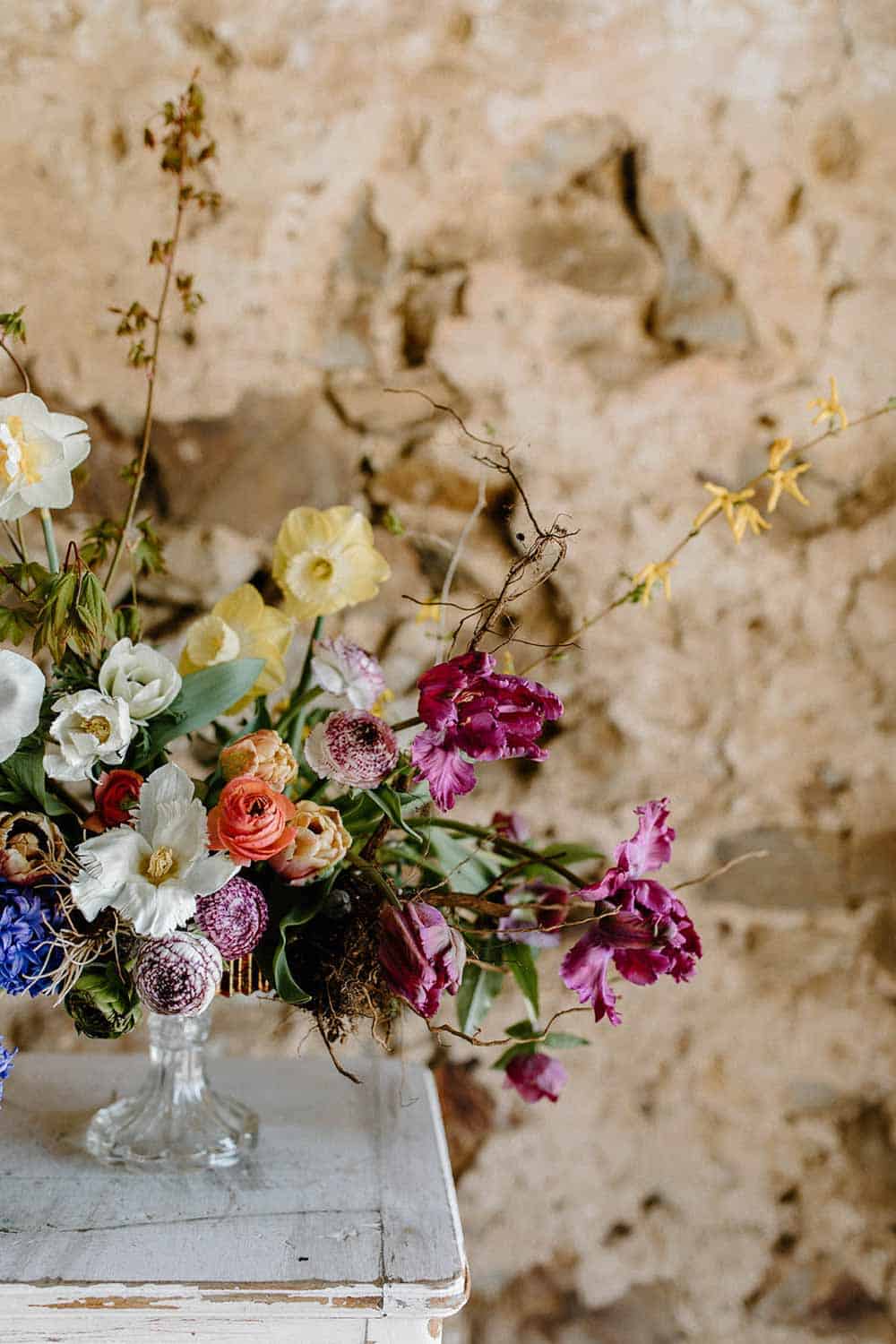 A floral arrangement with pink, purple, yellow, and white flowers in a glass vase on a white table against a rough, beige wall.