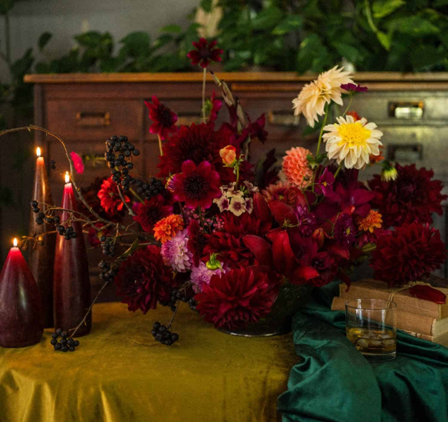 A vibrant floral arrangement in red and pink on a table with a drink, an open book, and three lit candles.