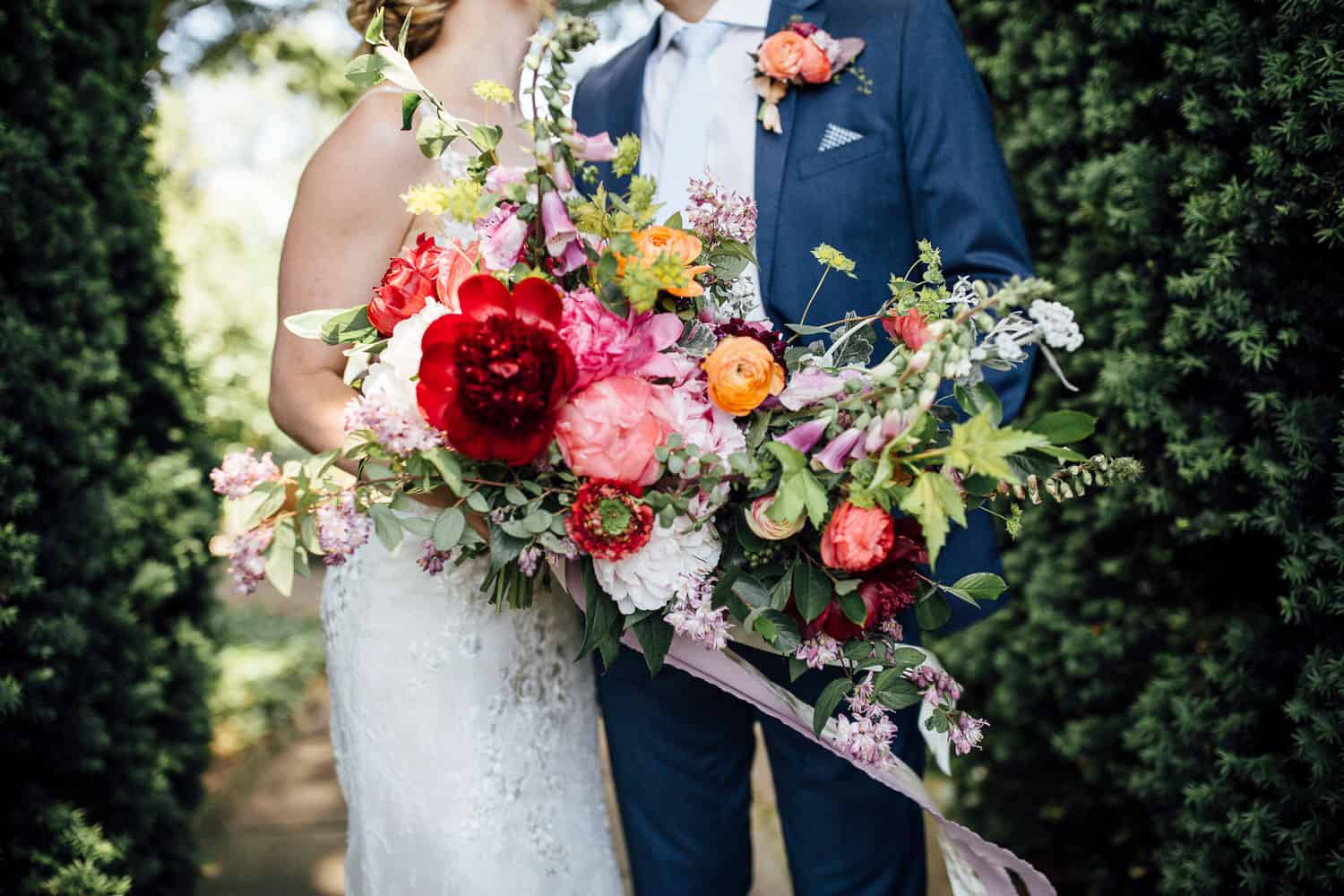 A bride in a white dress and a groom in a blue suit stand close together, holding a large, colorful bouquet of flowers.