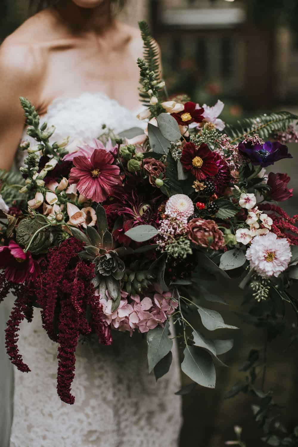 A woman in a white lace dress holds a large, colorful bouquet of various flowers and greenery.