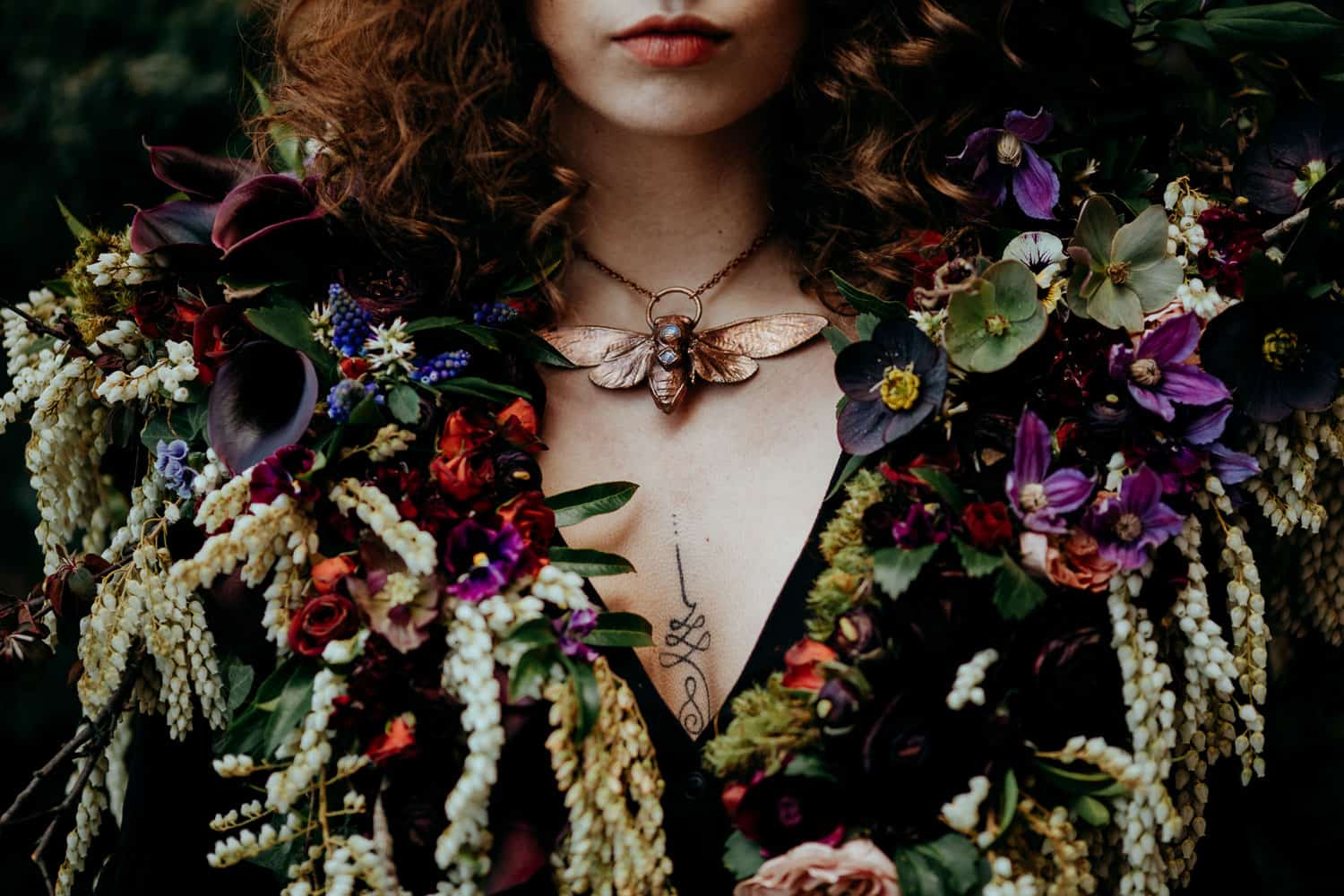 A woman with curly hair wears a necklace featuring a large insect pendant surrounded by various colorful flowers and plants.