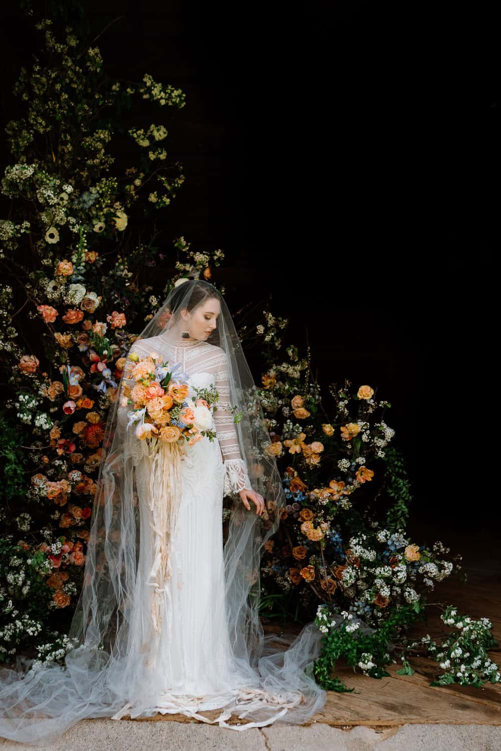 A bride in a white dress and veil holds a bouquet of orange, yellow, and white flowers, in front of a large floral arrangement.