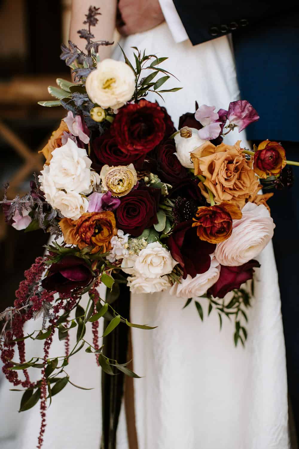 Close-up of a wedding bouquet held by a bride, with white, burgundy, and orange roses, greenery, and smaller flowers.