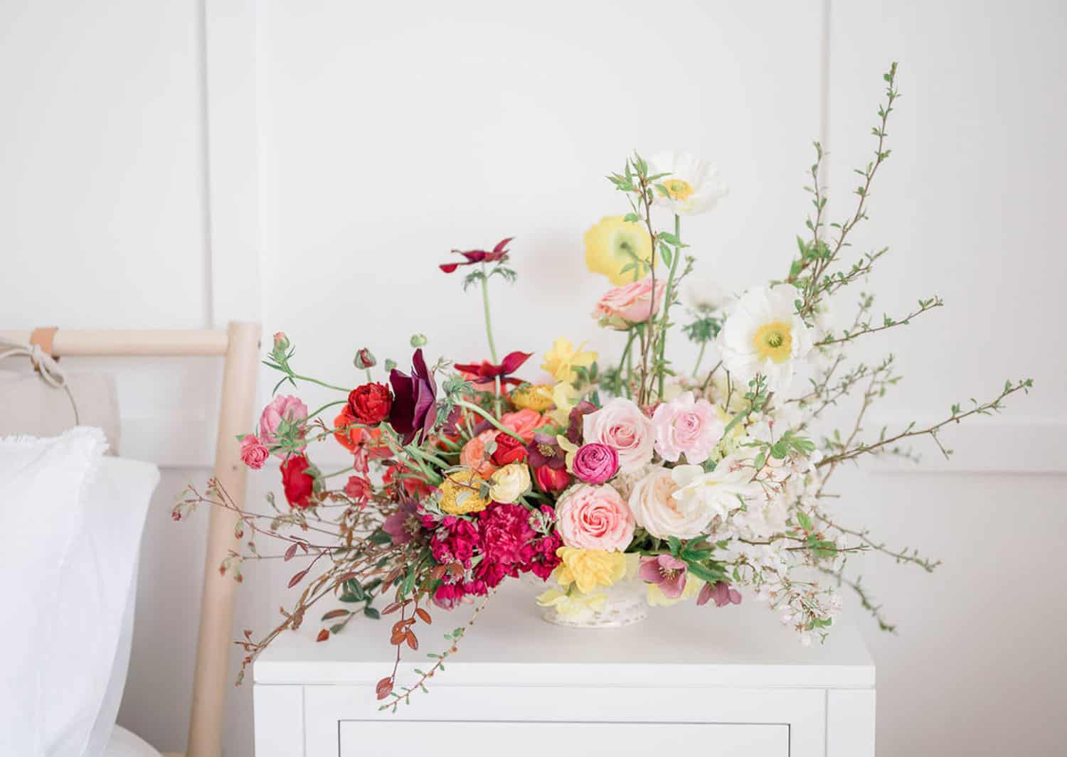 A floral arrangement with various colorful flowers, including roses, displayed on a white table against a white wall.