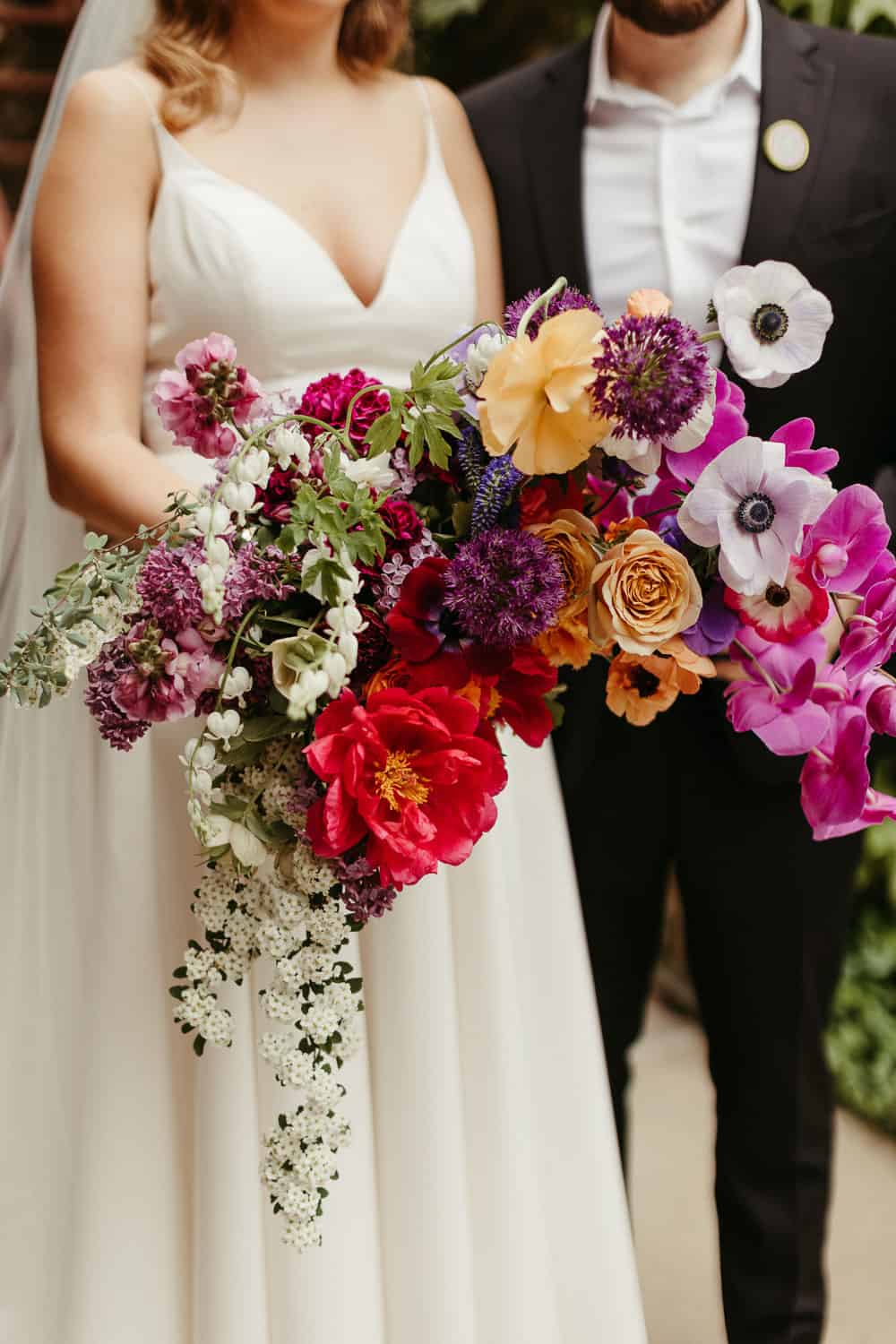 A bride and groom stand side by side with the bride holding a large colorful bouquet featuring red, purple, and pink blooms.