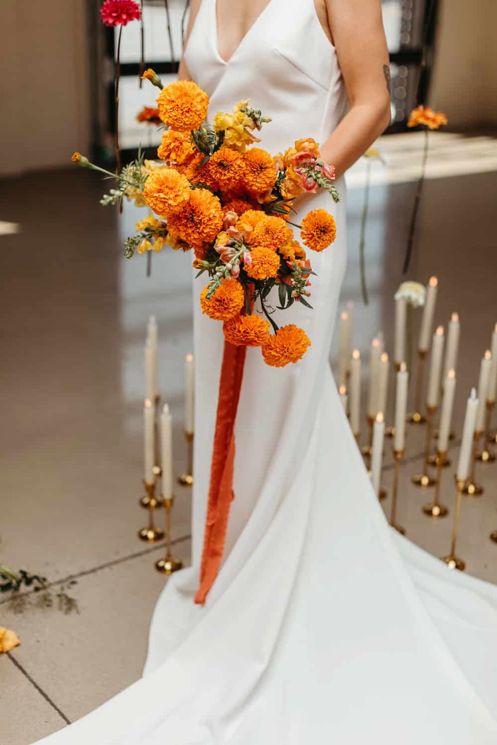 A bride in a white dress holds a bouquet of orange flowers, standing among multiple lit white candles arranged on the floor.