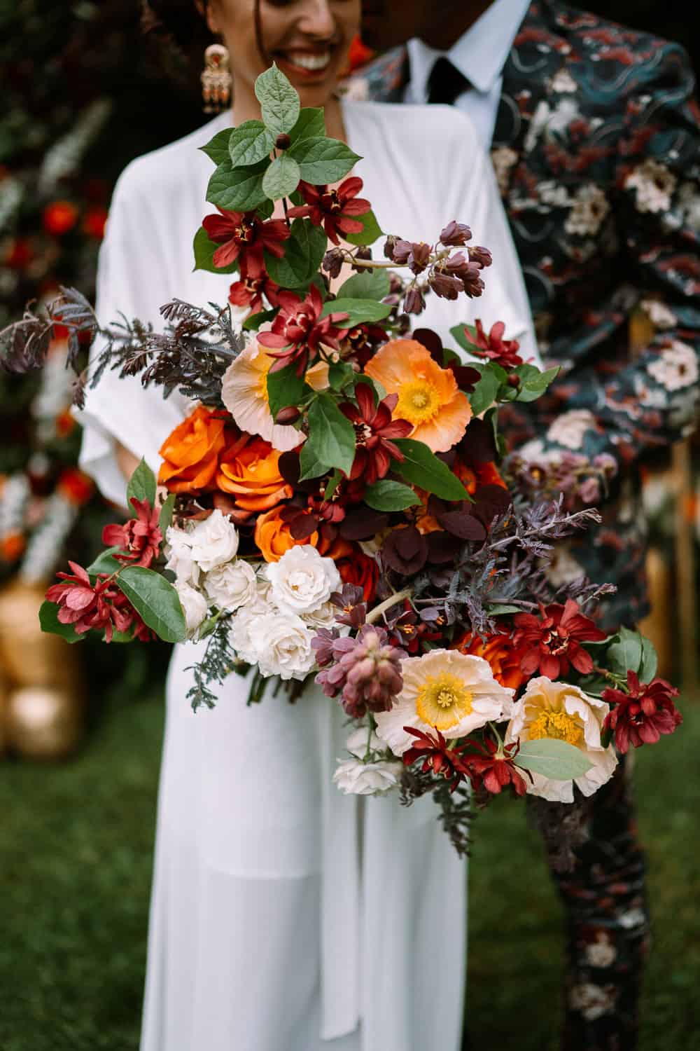 A woman in a white dress holds a large, vibrant bouquet of flowers, standing beside another person in a suit.