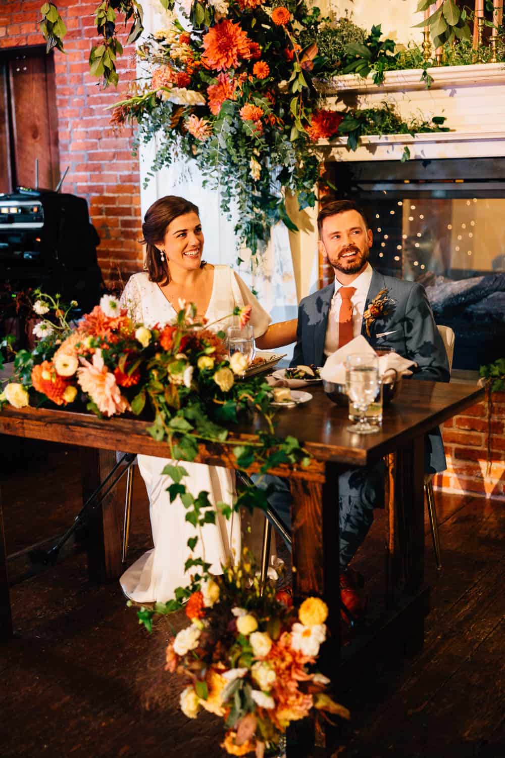 A bride and groom sit at a decorated table in a room with brick walls and a fireplace adorned with floral arrangements.