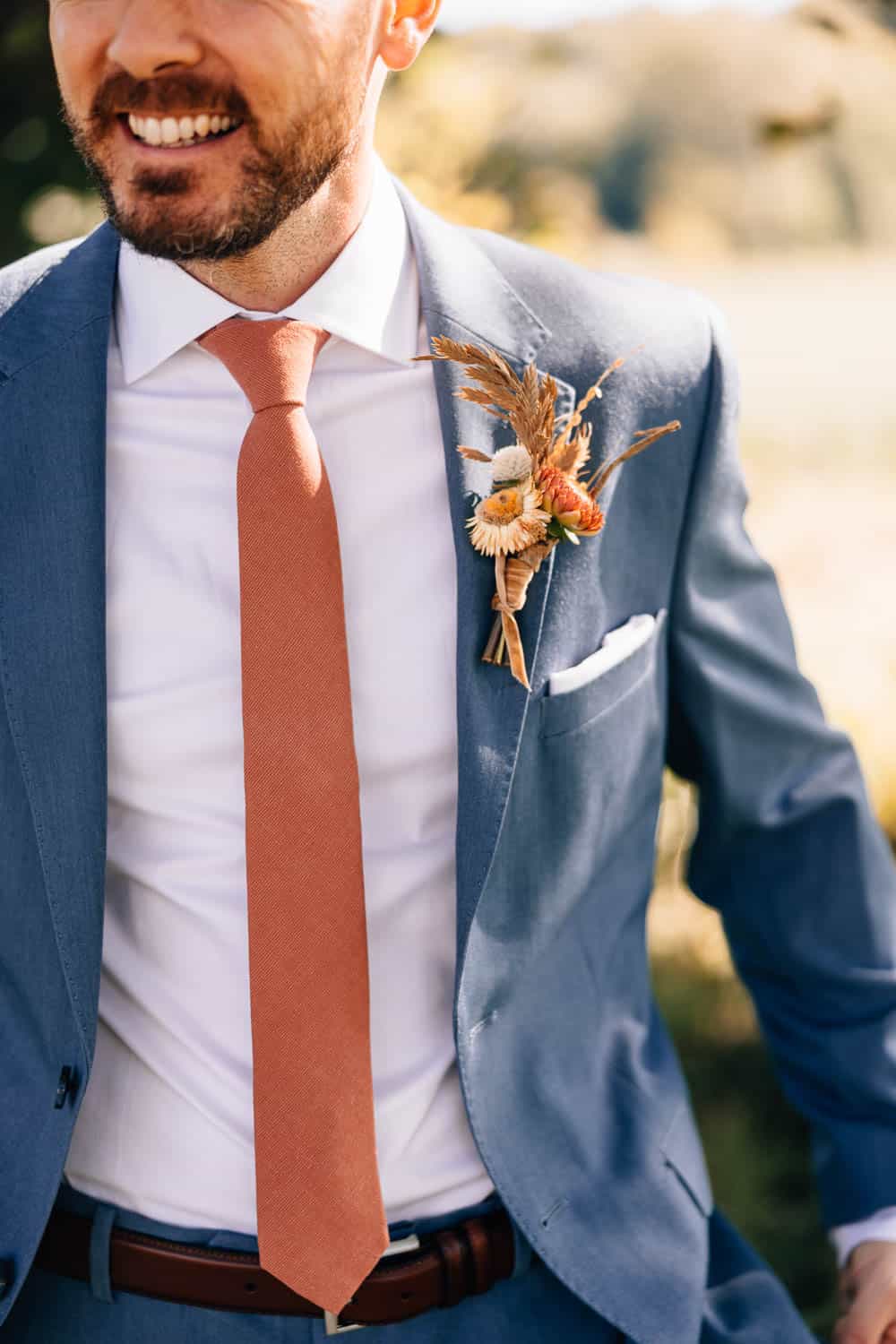 A man in a blue suit and rust-colored tie with a boutonnière smiles outdoors.