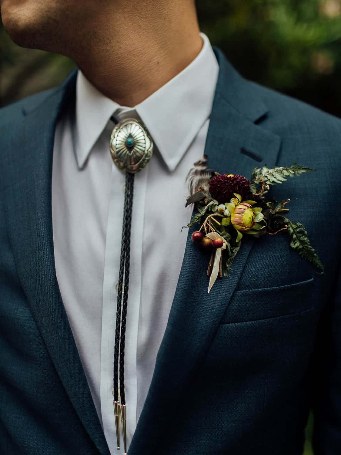 Close-up of a person wearing a dark blue suit with a white shirt, a bolo tie, and a floral boutonnière.