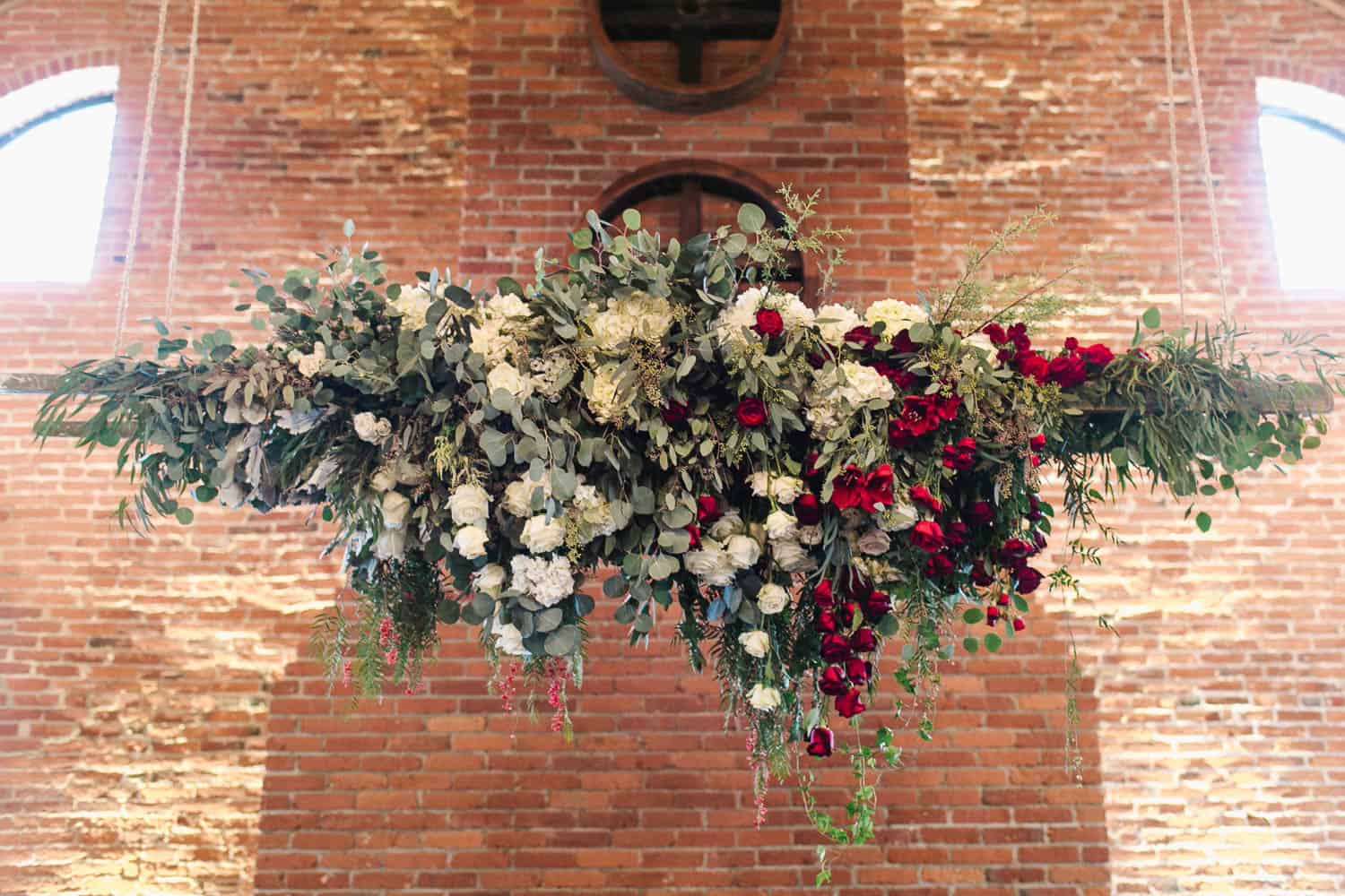 A floral arrangement featuring a mix of white and red flowers with green foliage, hanging against a brick wall backdrop.